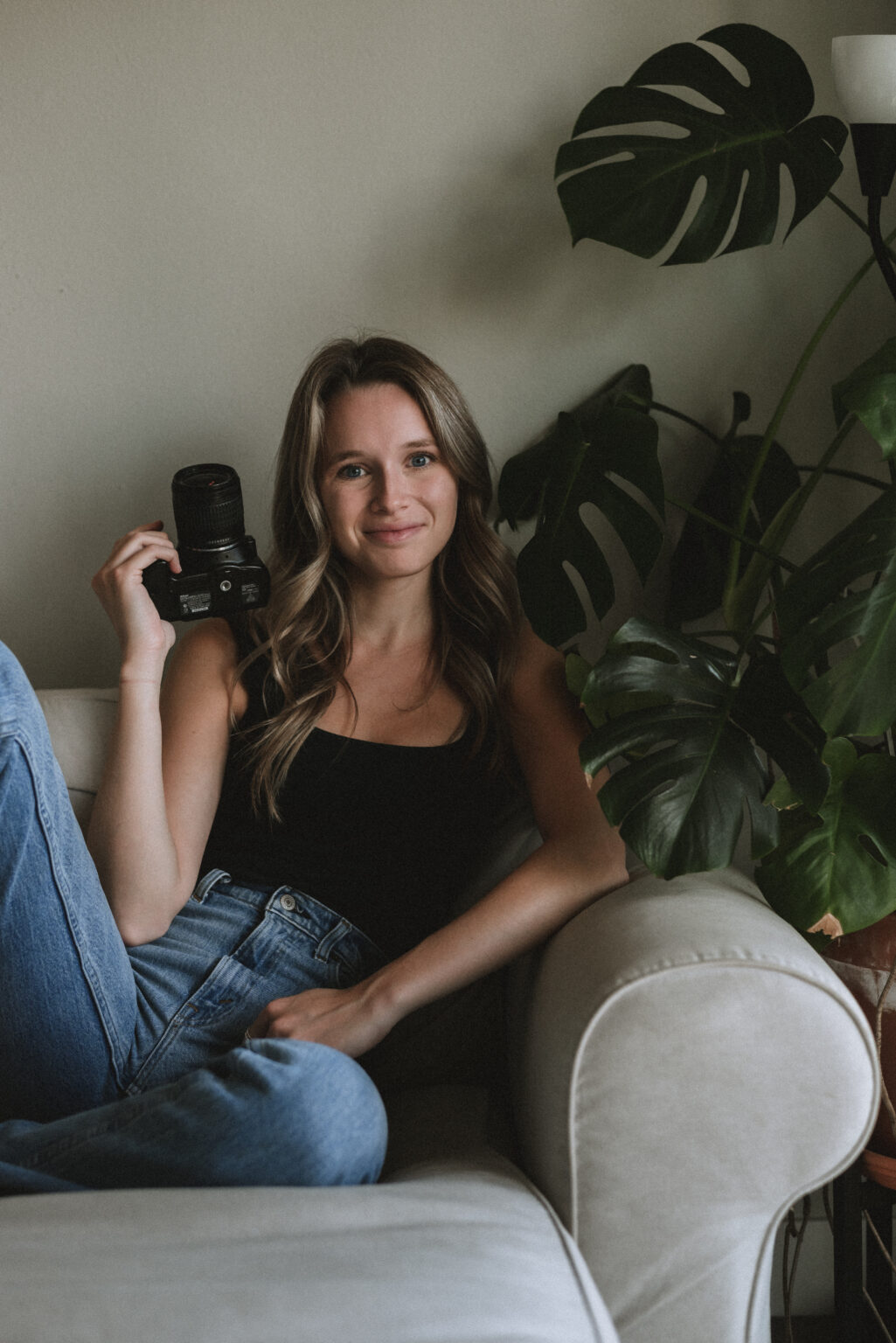 Julia Johnson soft smiling with camera in hand while sitting on a white/sand colored couch. There is a Monstera plant next to her.